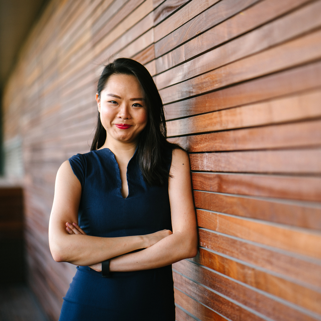 Woman in blue dress with arms crossed, smiling against a wooden wall background, conveying a confident and friendly mood.