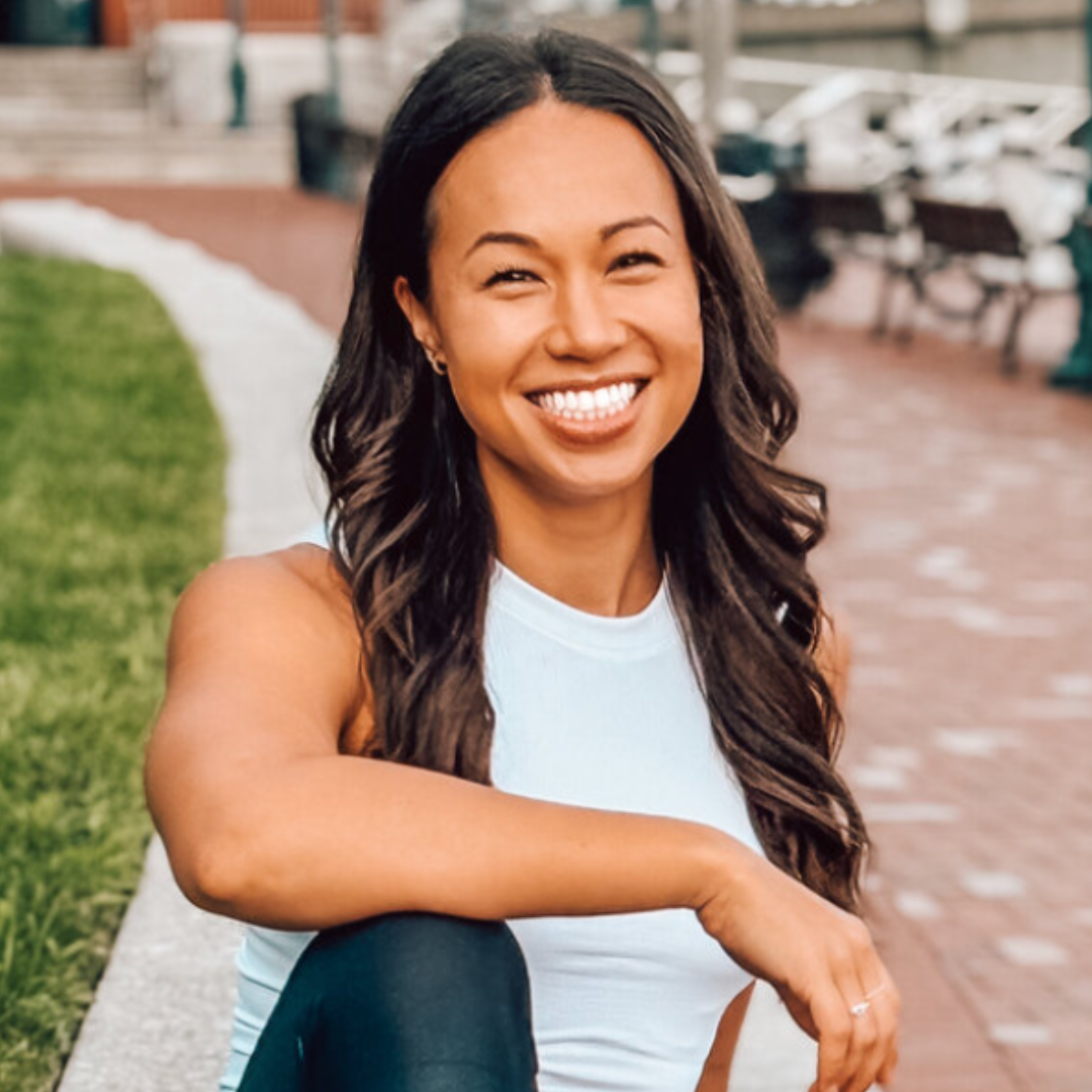 A woman in a light blue top smiles while sitting in an outdoor park with brick paths and green grass, creating a cheerful, sunny atmosphere.