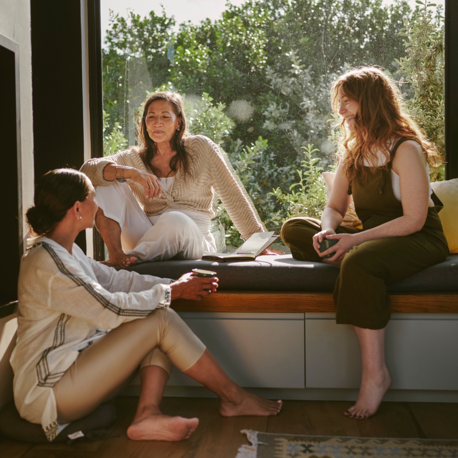 Three women sit by a window with green plants outside, talking and holding mugs. They appear relaxed and engaged. Sunlight filters in.