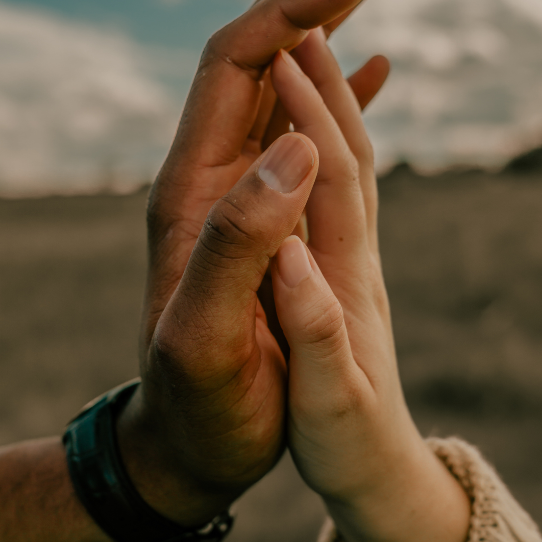 Two hands gently touching, set against a blurred outdoor background with cloudy sky. A watch is visible on one wrist. Peaceful mood.