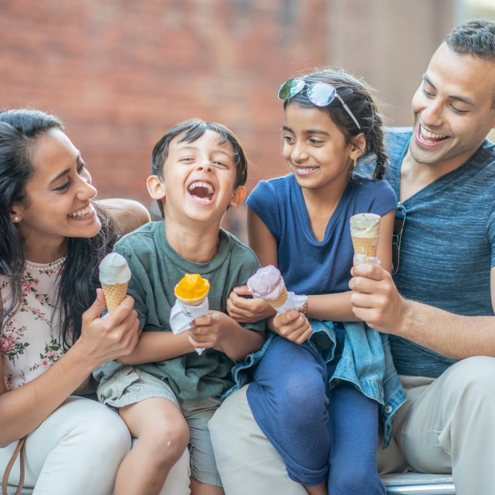 Family of four smiles while eating colorful ice cream cones outdoors, with a brick wall in the background. Fun, joyful mood.