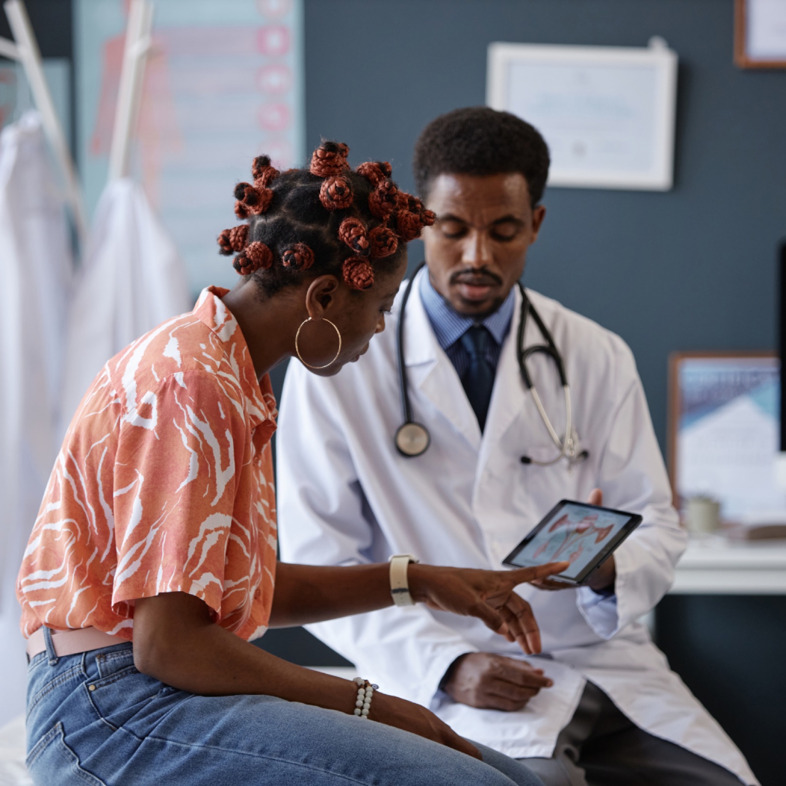 Doctor and patient discussing on a tablet in a clinic. Patient wears a patterned shirt, doctor in a white coat. Informative and calm setting.
