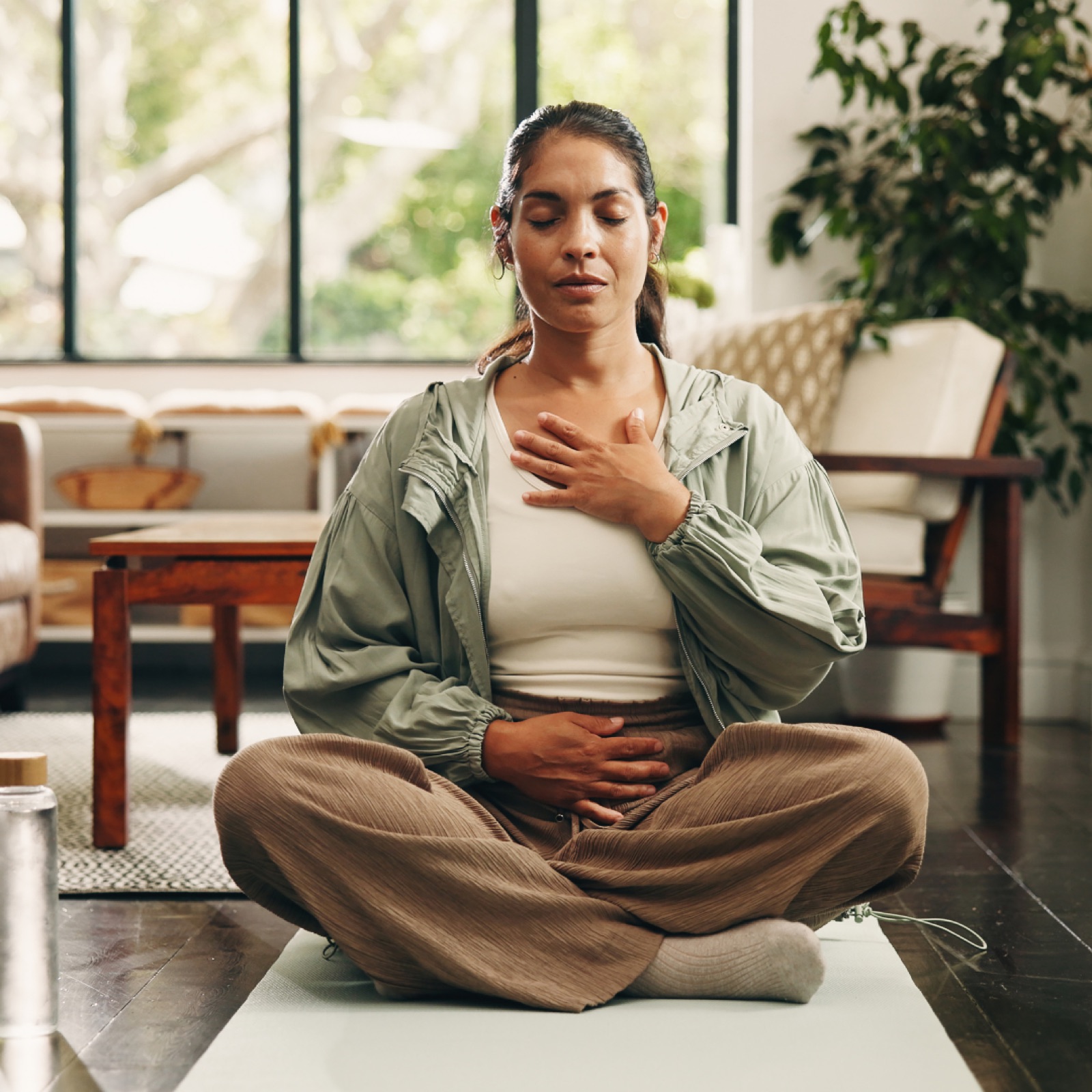Woman meditating indoors, seated cross-legged on a mat with eyes closed, hands on chest and stomach. Relaxed mood, plants in background.
