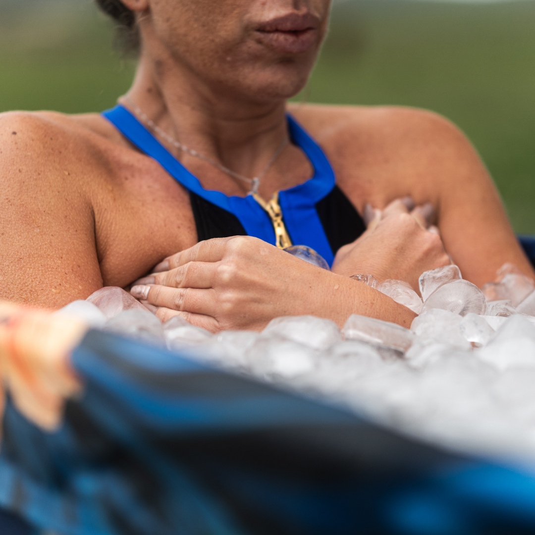 Woman in a blue swimsuit sits in an ice bath, arms crossed over chest. Calm expression, blurred green background, and focus on ice.