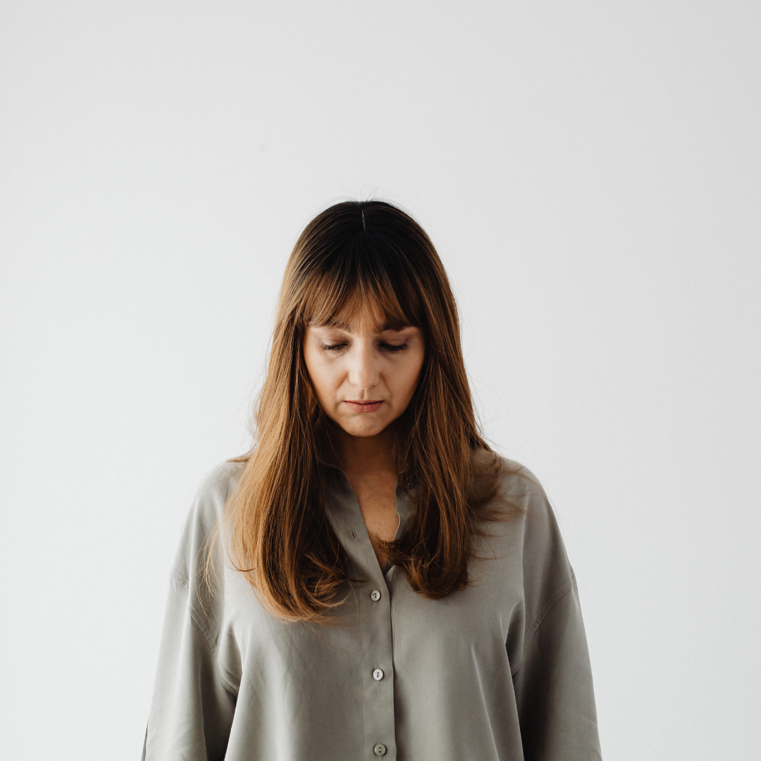 Woman with long brown hair in a gray shirt looks down, standing against a plain white background. Sad expression, minimalist setting.