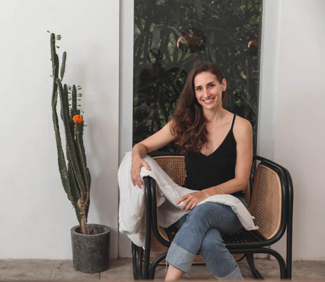 Woman in black top sits smiling on a wicker chair by a tall cactus in a pot. Background shows lush greenery through a glass window.