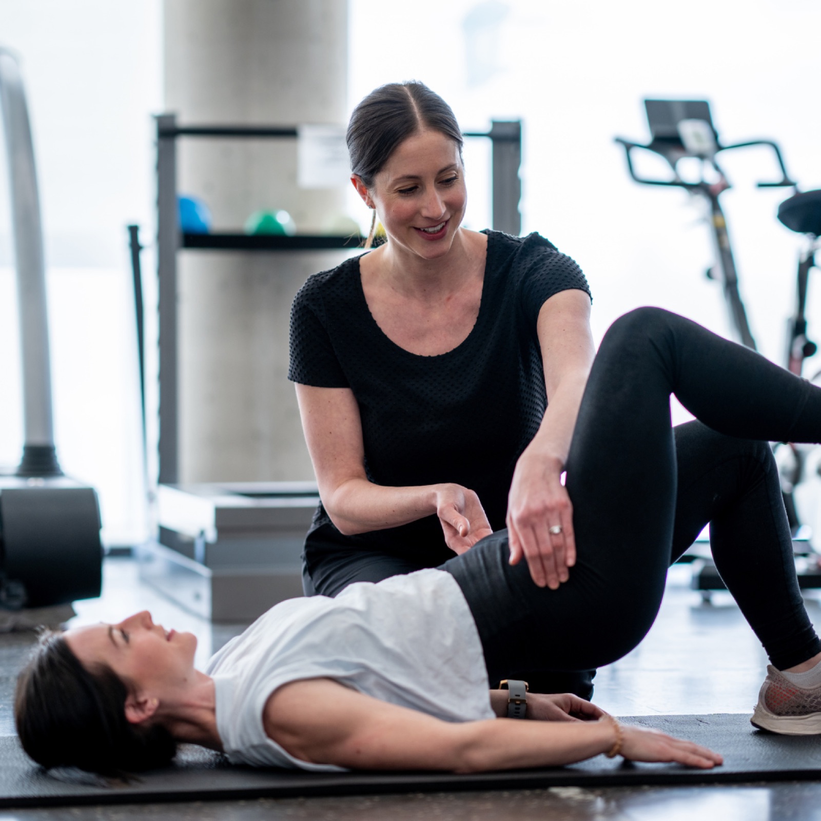 Trainer assists woman with exercise on mat in gym. Trainer smiles, wearing black. Background has gym equipment and bright lighting.