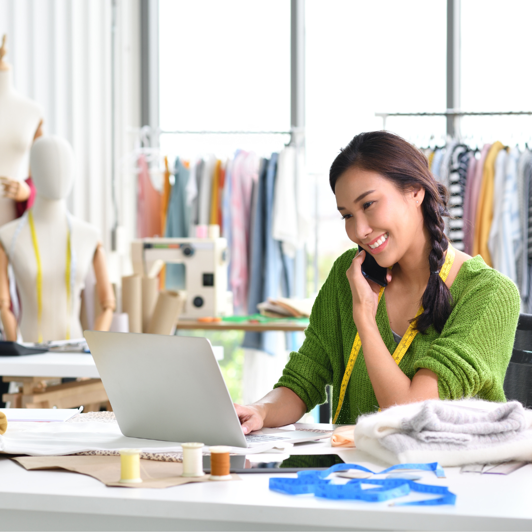 Woman in green sweater on phone, smiling at laptop in sewing studio. Mannequin, sewing machine, fabrics, and clothes rack in background.
