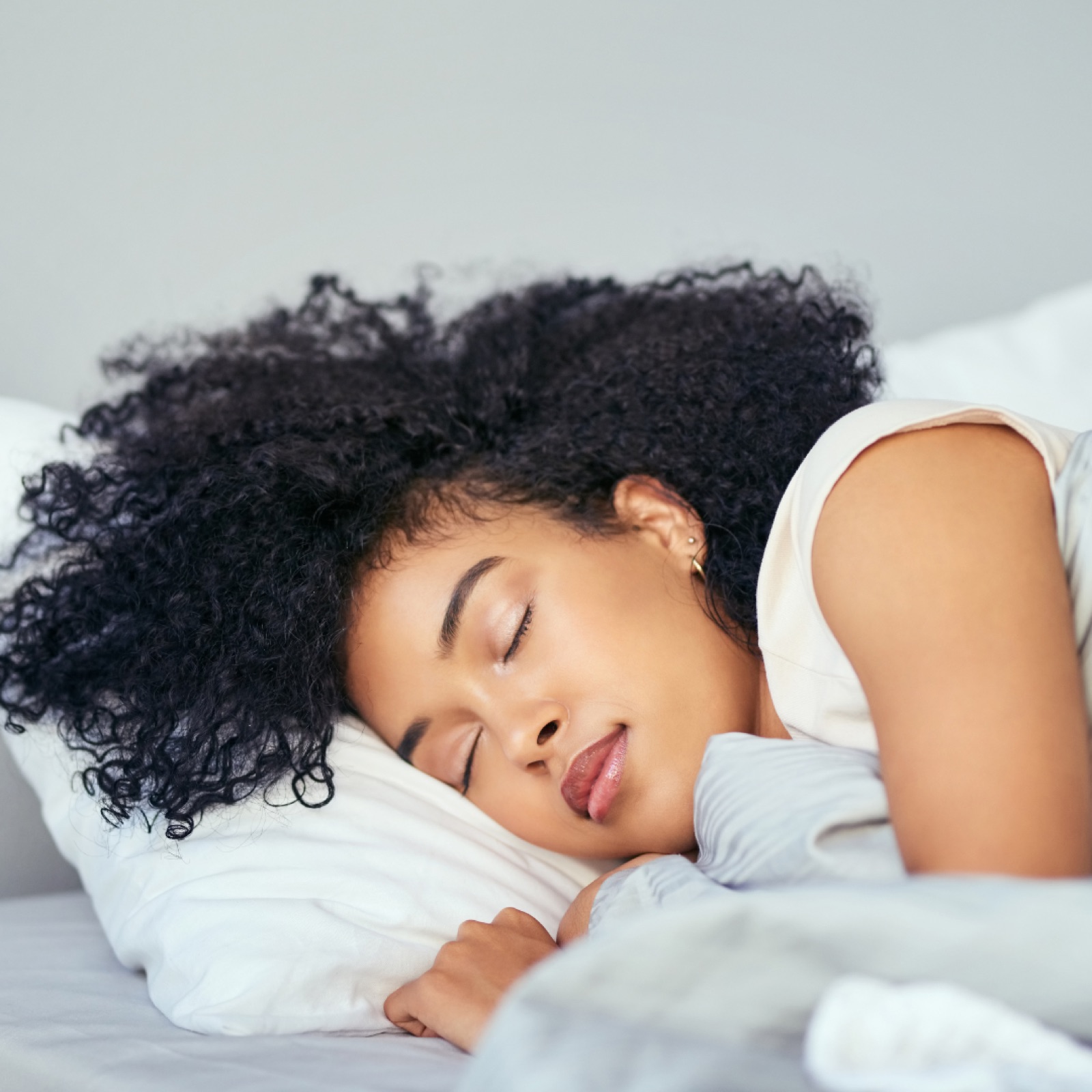 A person with curly hair sleeps peacefully on a white pillow in a bright room, giving a serene and restful impression.