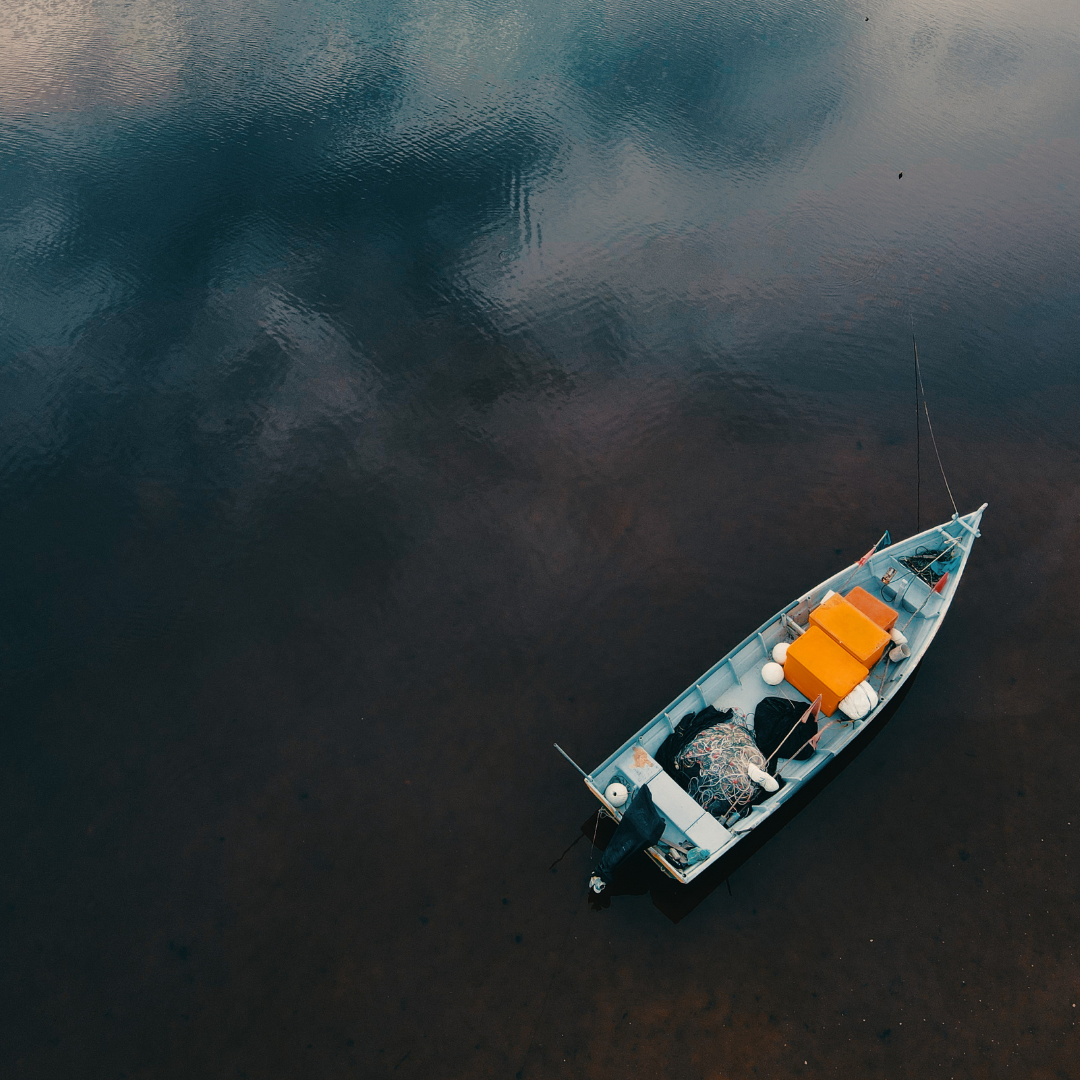 A small boat with fishing nets and orange seats floats on dark, reflective water under a cloudy sky, creating a calm, serene mood.
