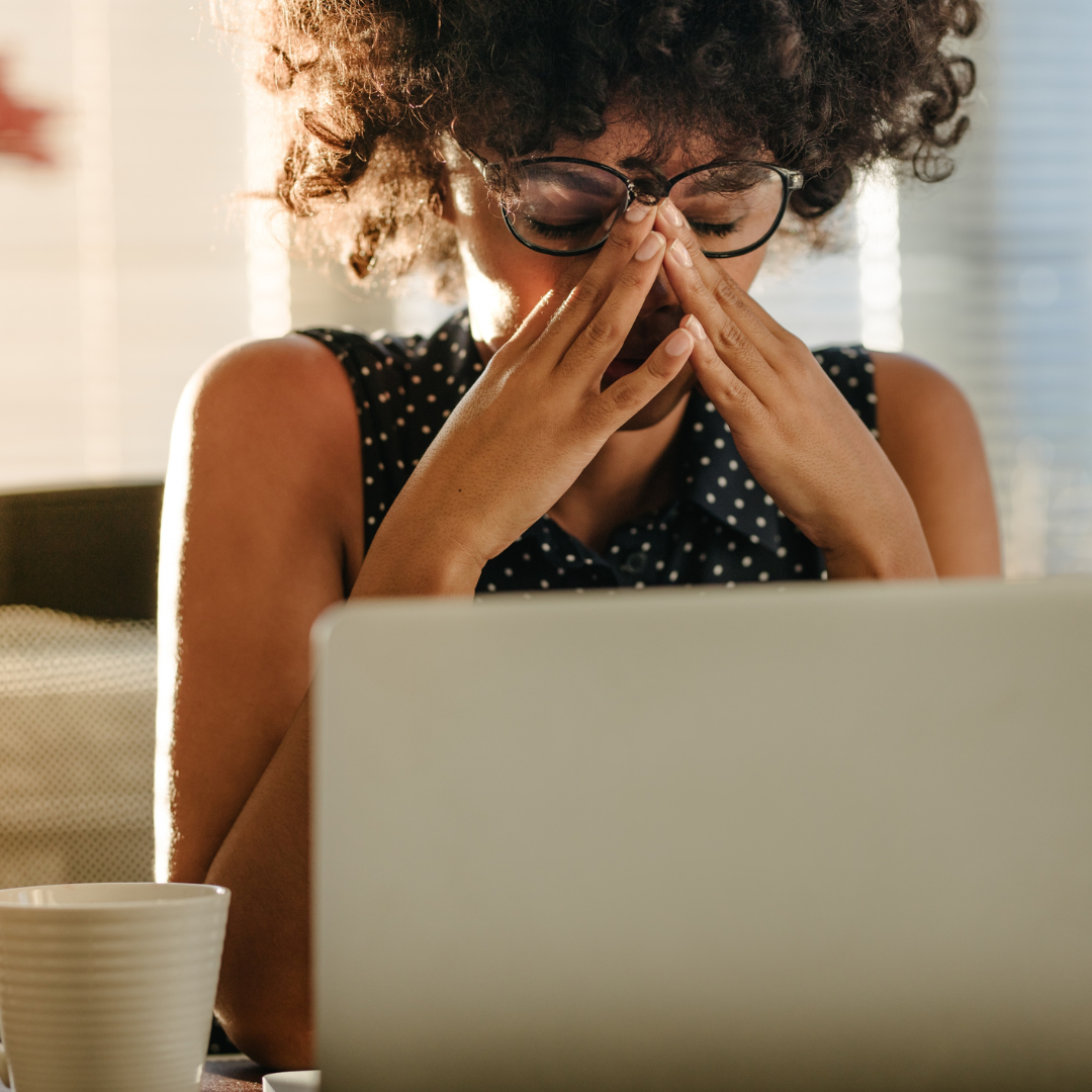 A woman in glasses, appearing stressed, sits at a desk with a laptop in front of her, rubbing her eyes. Sunlight streams through blinds.