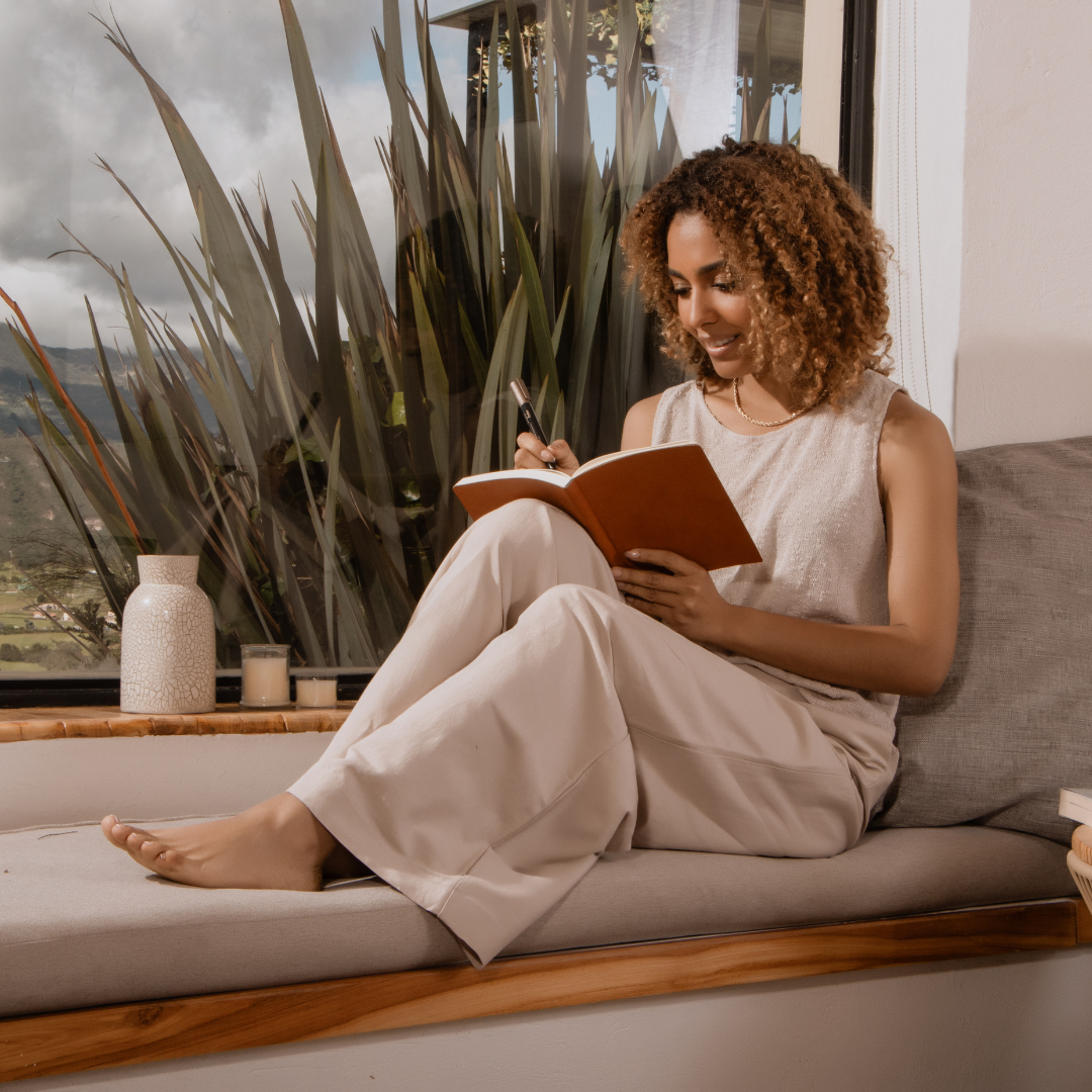 Woman with curly hair writes in a notebook, sitting by a window with plants outside. She's on a cushioned bench, relaxed and content.