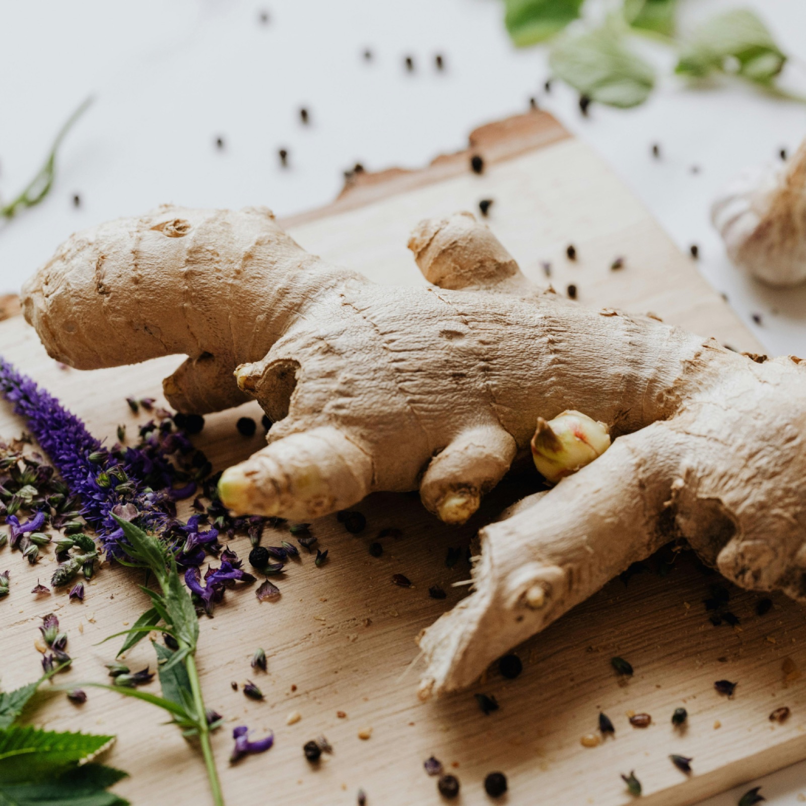 Ginger root on a wooden board with purple flowers and peppercorns scattered around. Fresh green leaves in the background. Warm, earthy tones.