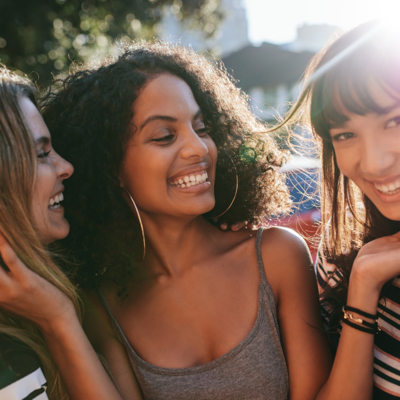 Three women laughing together outdoors, sunlit background. They wear casual clothing, with one in hoop earrings. Joyful and candid moment.