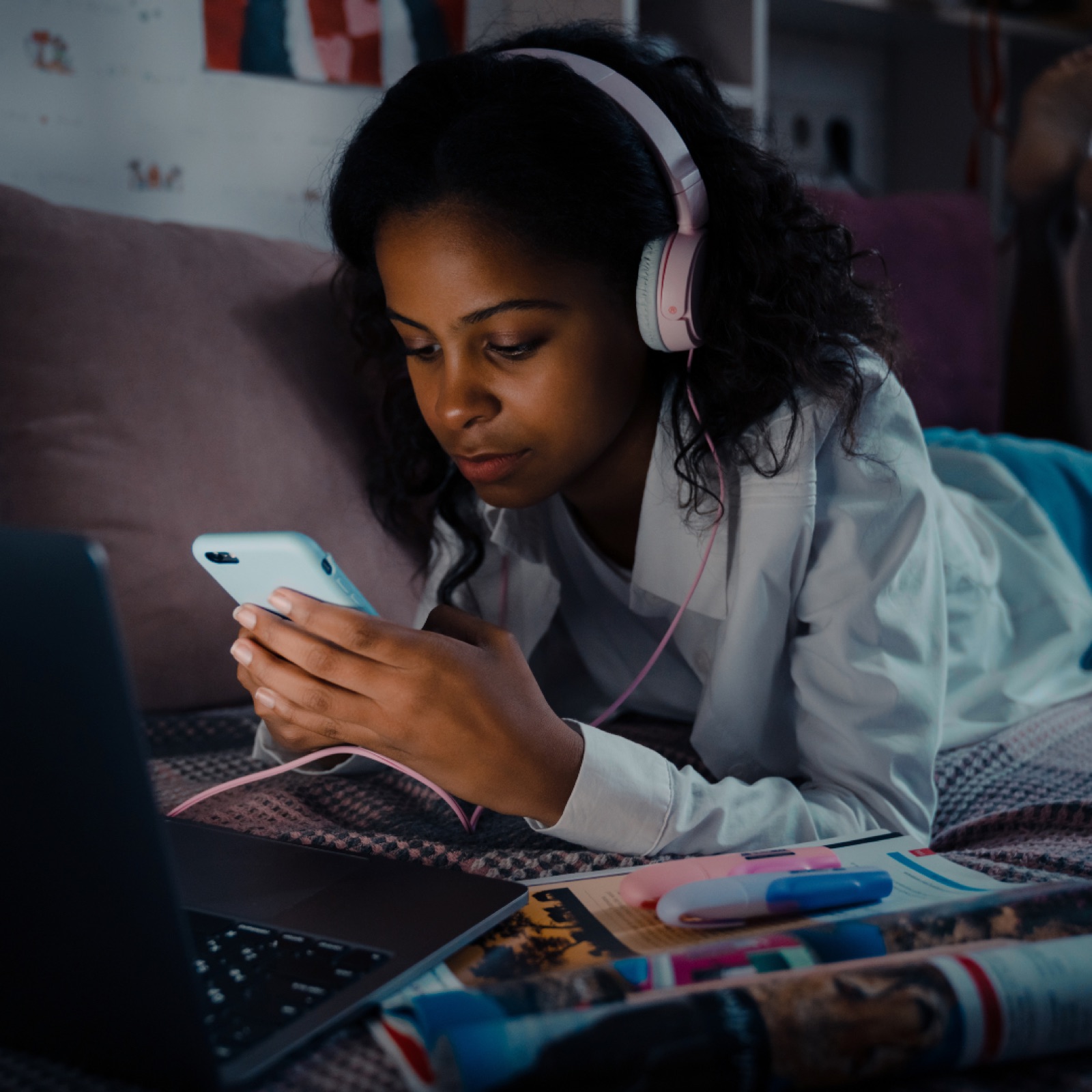 Young woman with headphones focused on smartphone, lying on bed. Nearby are an open laptop, magazines, and colorful markers. Dimly lit room.