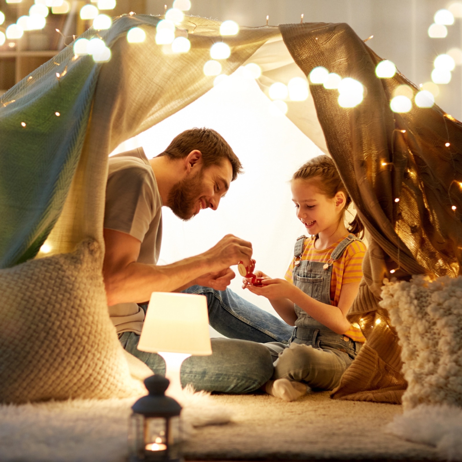 Father and daughter in a cozy blanket fort with string lights, playing joyfully with toys. Warm lighting and soft décor create a playful mood.