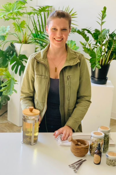 Smiling person in green jacket stands at table with tea, jars, and cutlery. Lush plants in the background create a fresh atmosphere.