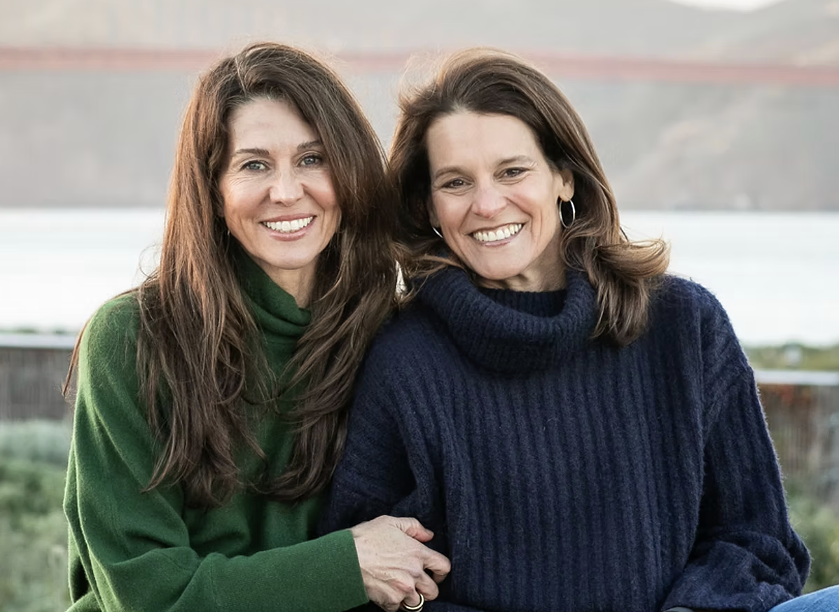 Two smiling women in green and navy sweaters pose outdoors. A blurred bridge is in the background, suggesting a calm and happy mood.
