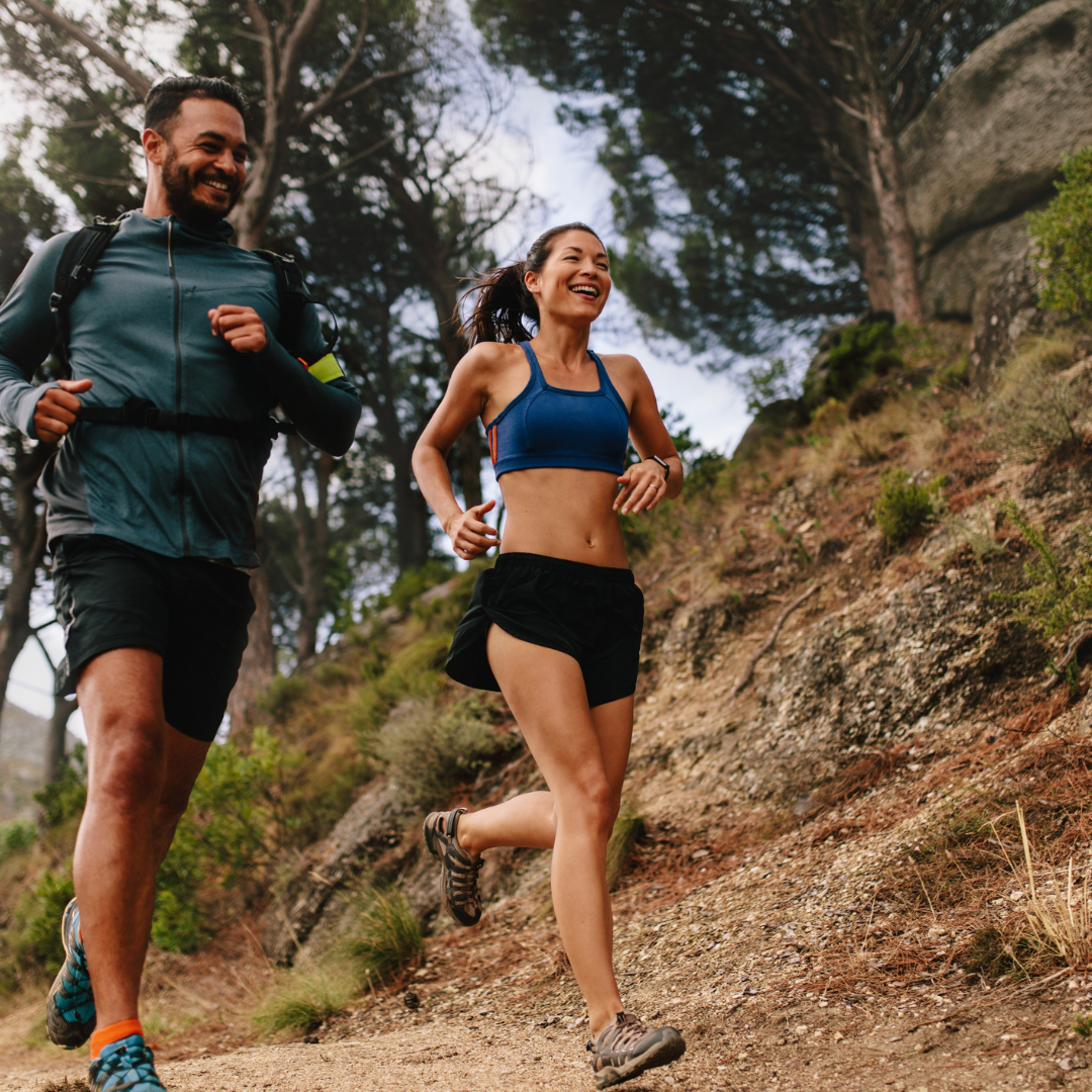 Two people joyfully trail running on a forest path. The man wears a jacket; the woman, a blue sports bra. Lush greenery and trees surround them.