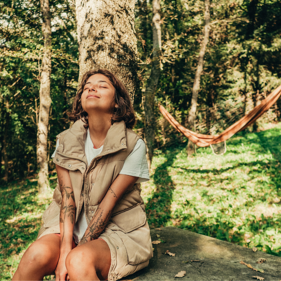 Woman in beige vest sitting against a tree, eyes closed, smiling serenely. Sunlit forest background with hammock and green grass.