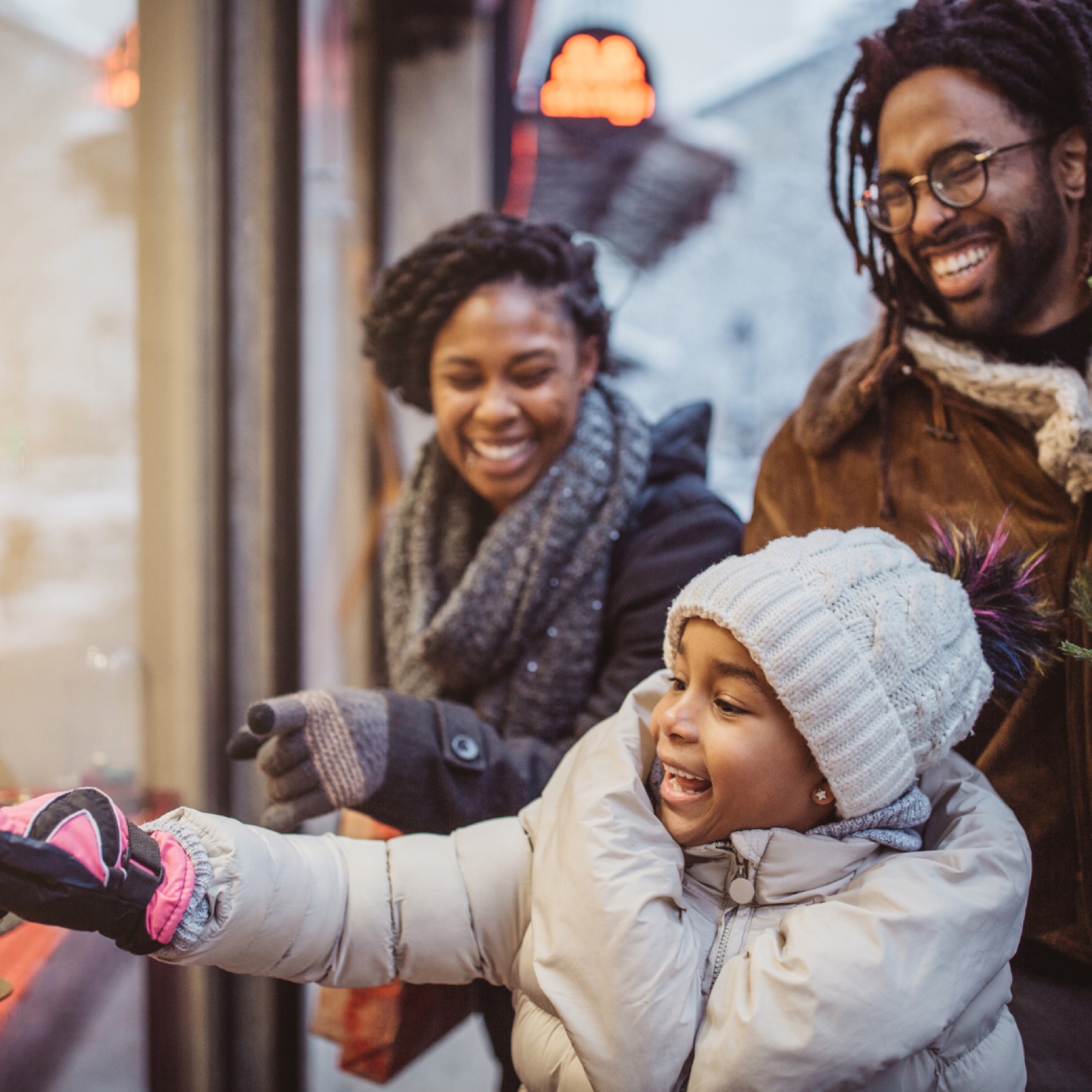 A family of three, smiling and dressed warmly, looks at a shop window. The child, in a white beanie, excitedly points inside. Snowy background.