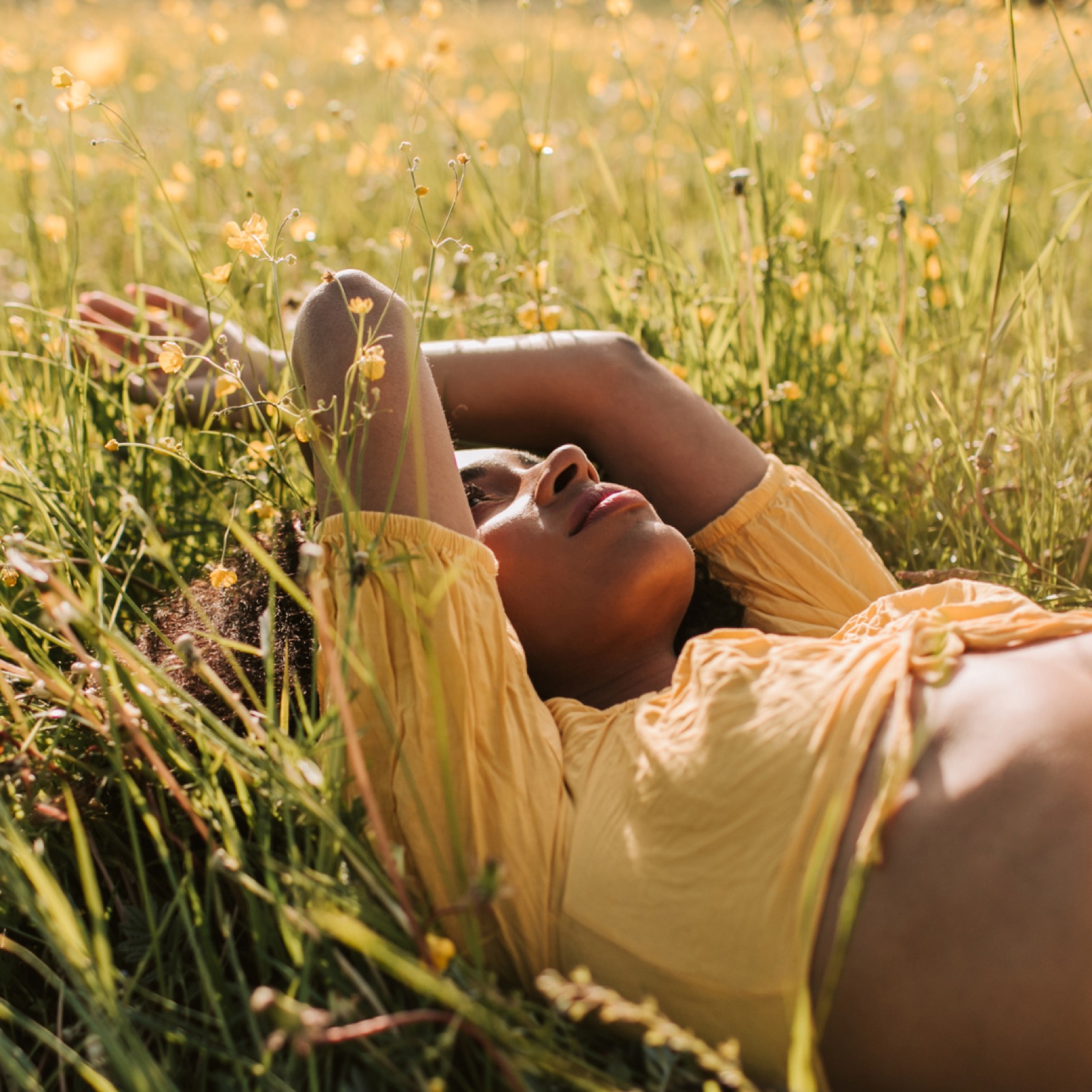 Woman in a yellow dress lying peacefully in a field of yellow flowers, arms above her head, under sunny skies, conveying tranquility.