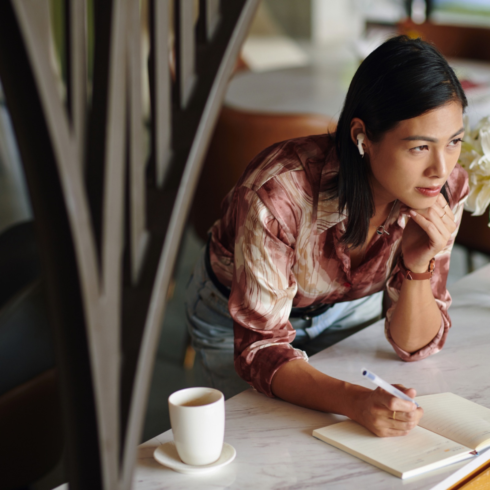 Woman in patterned shirt, wearing earbuds, leans on cafe table, writing in a notebook. A coffee cup sits nearby. Calm atmosphere.