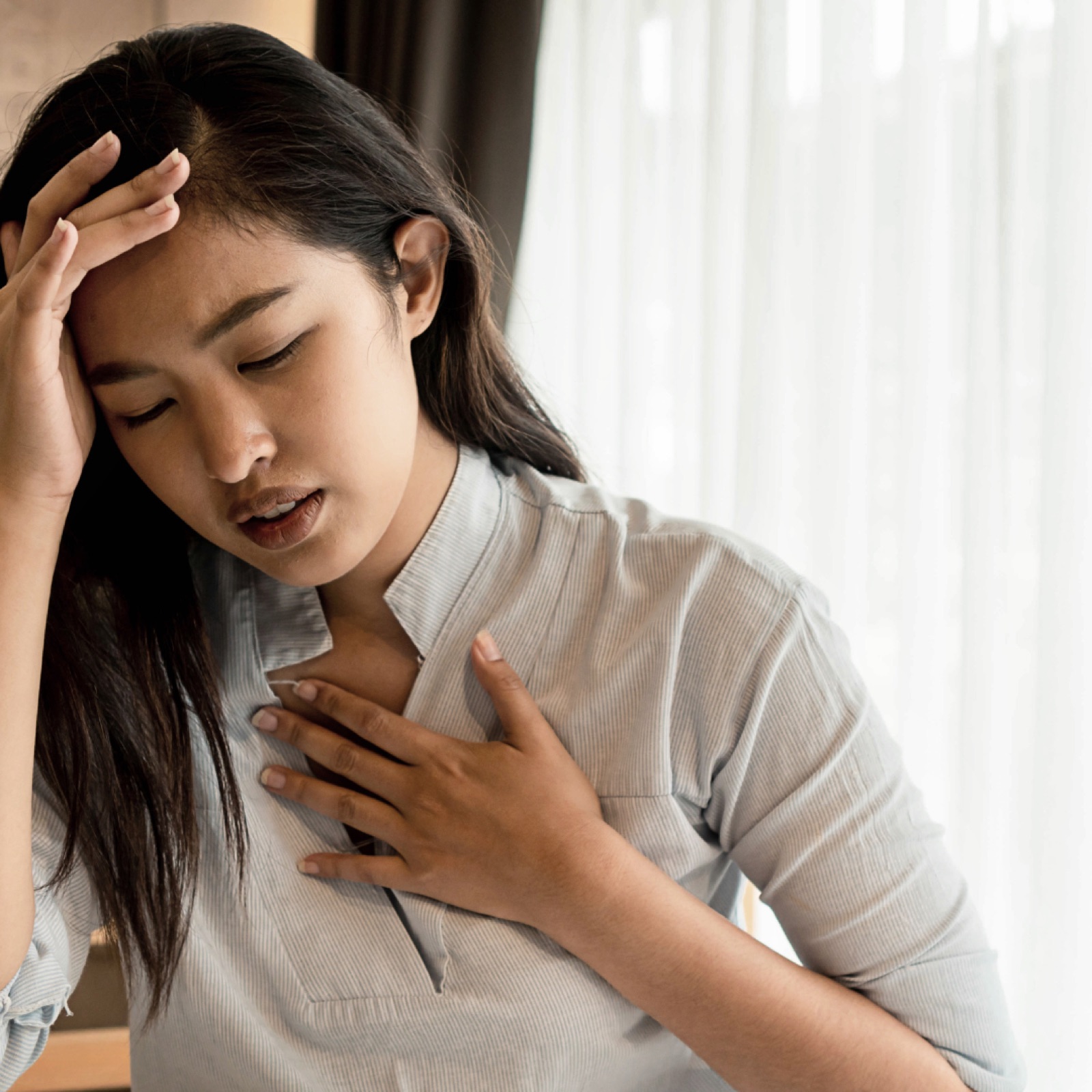 Woman in light gray shirt holds her head and chest, appearing distressed. She's indoors, with white curtains in the background.