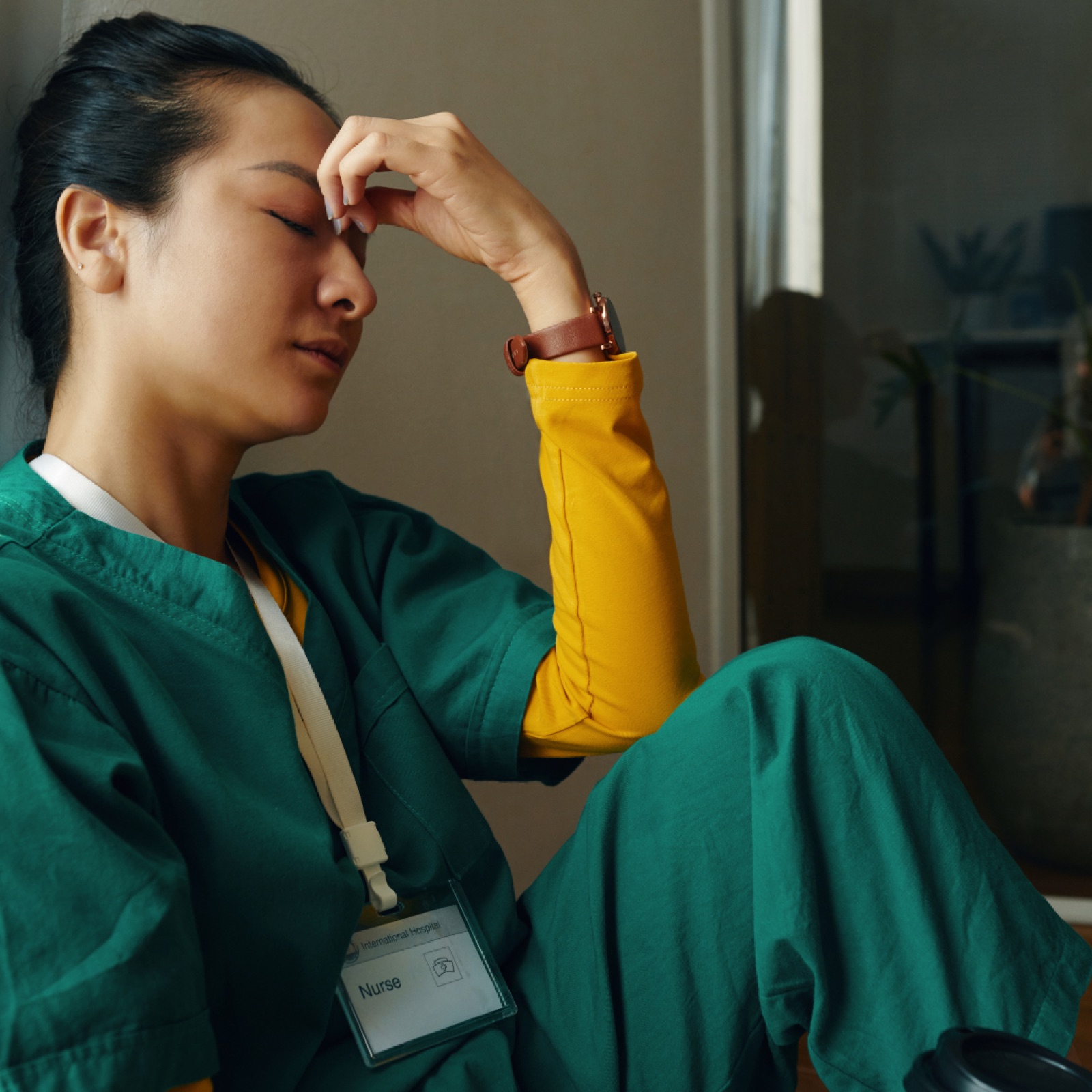 Nurse in green scrubs and yellow top sits with closed eyes, hand on forehead. Relaxed posture, muted room background. Name tag visible.