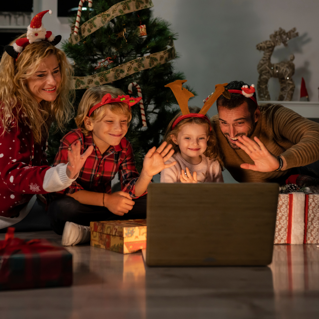 Family with festive headbands waves at a laptop in front of a decorated Christmas tree, surrounded by gifts. Warm and joyful atmosphere.