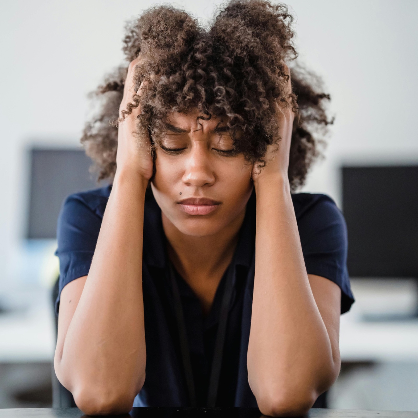 Woman with curly hair in blue shirt, sitting with hands on head, eyes closed, appears stressed. Office setting, blurred background.