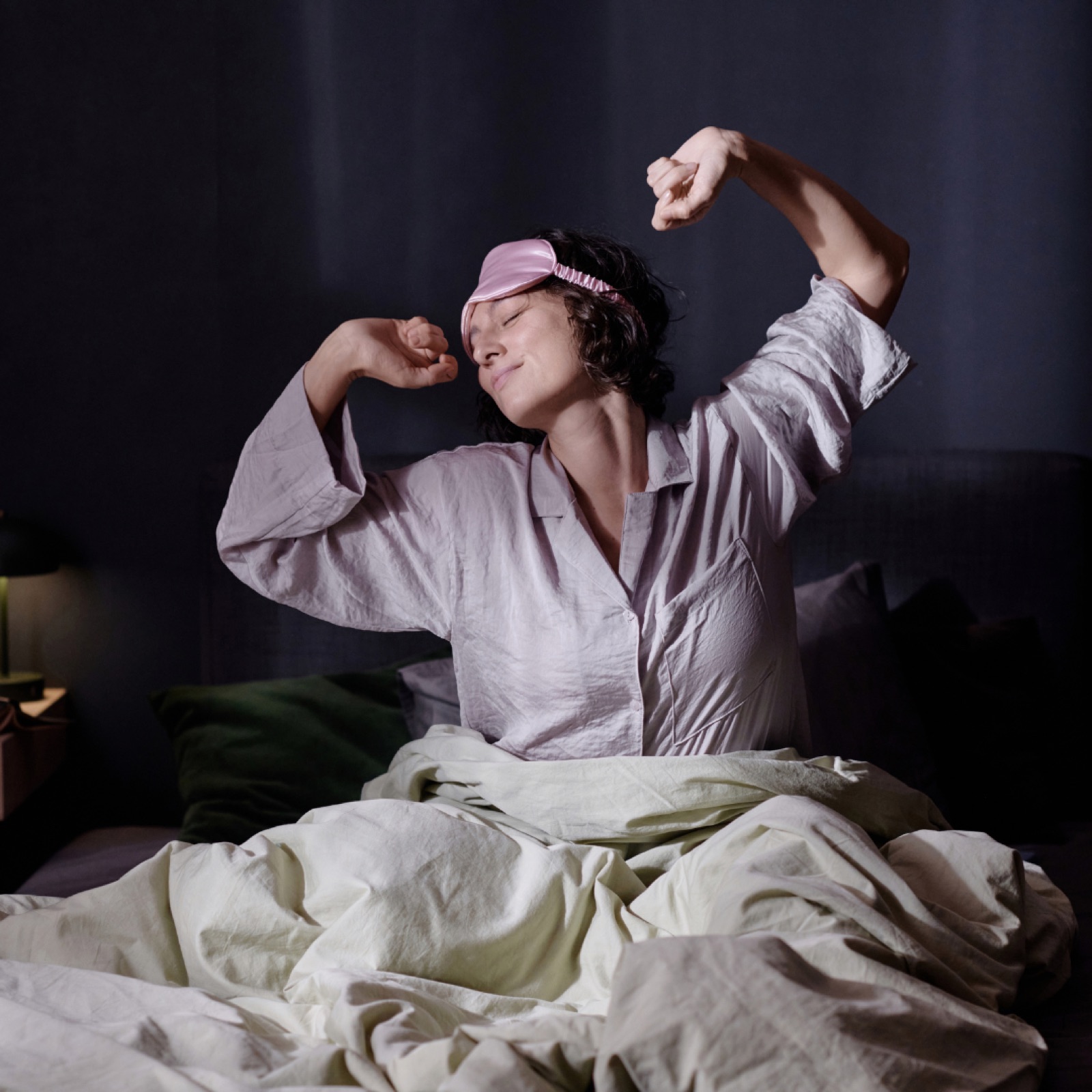 Woman in white pajamas and pink sleep mask stretching in bed, with cozy green and white bedding in a dimly lit bedroom.