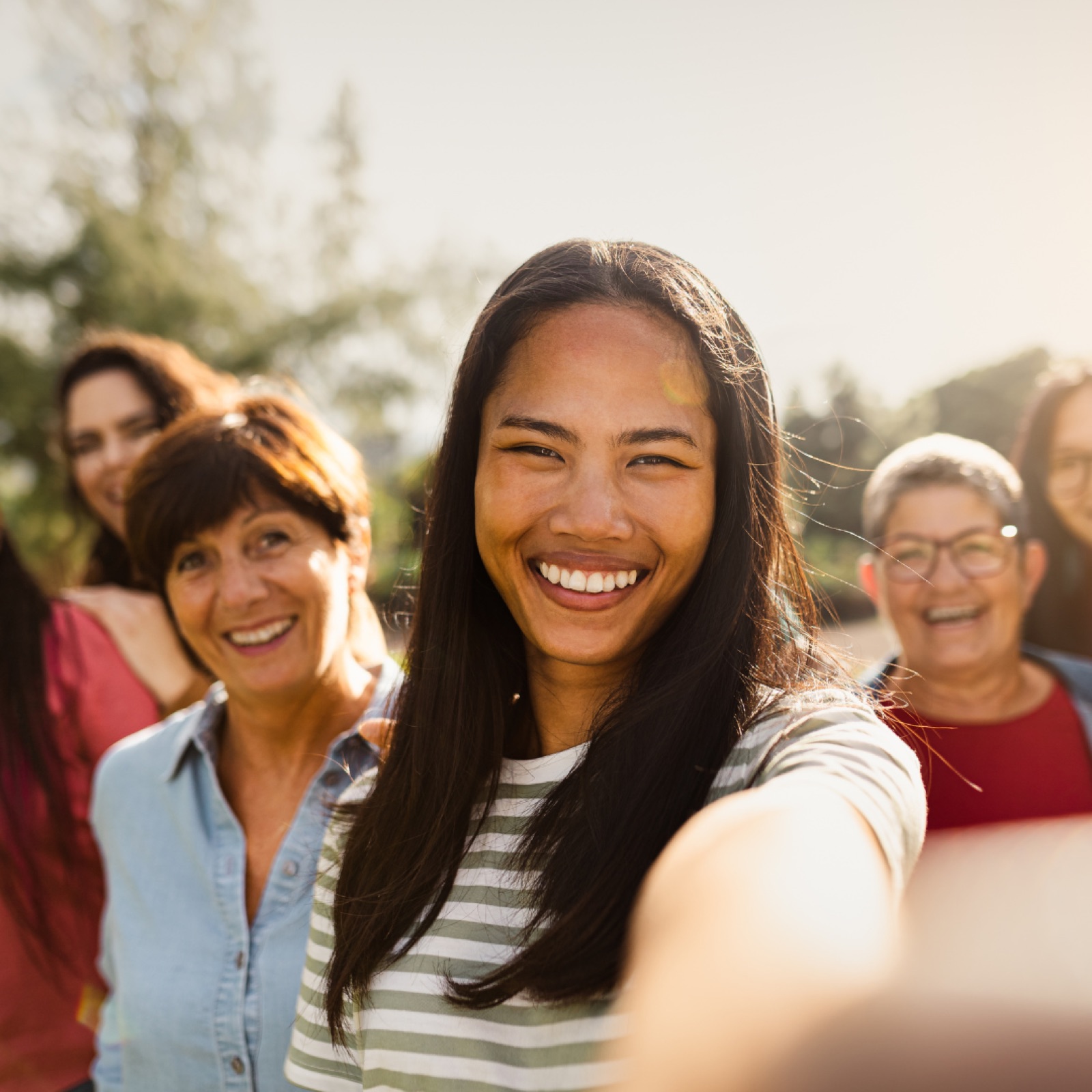 Smiling group selfie outdoors, diverse women enjoying sunlight. Casual attire, striped shirt in front, greenery in background, cheerful mood.