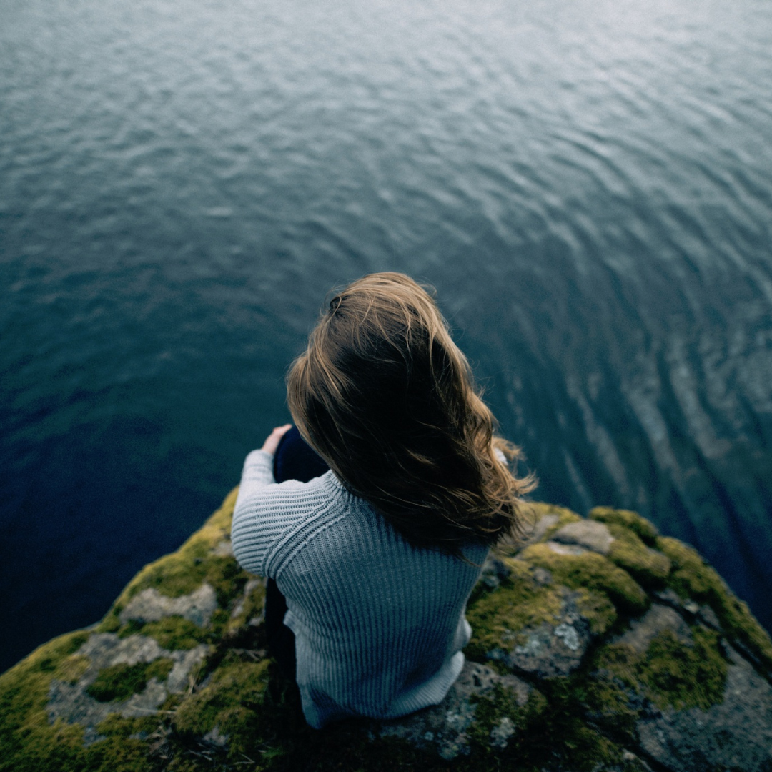 Person in a gray sweater sits on a mossy rock cliff overlooking calm water, conveying a peaceful, contemplative mood.