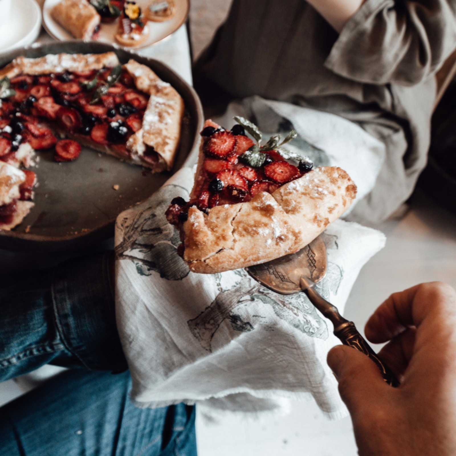 Hand holds a slice of berry galette on a patterned napkin. A larger galette sits on a plate nearby. Warm, inviting, rustic setting.