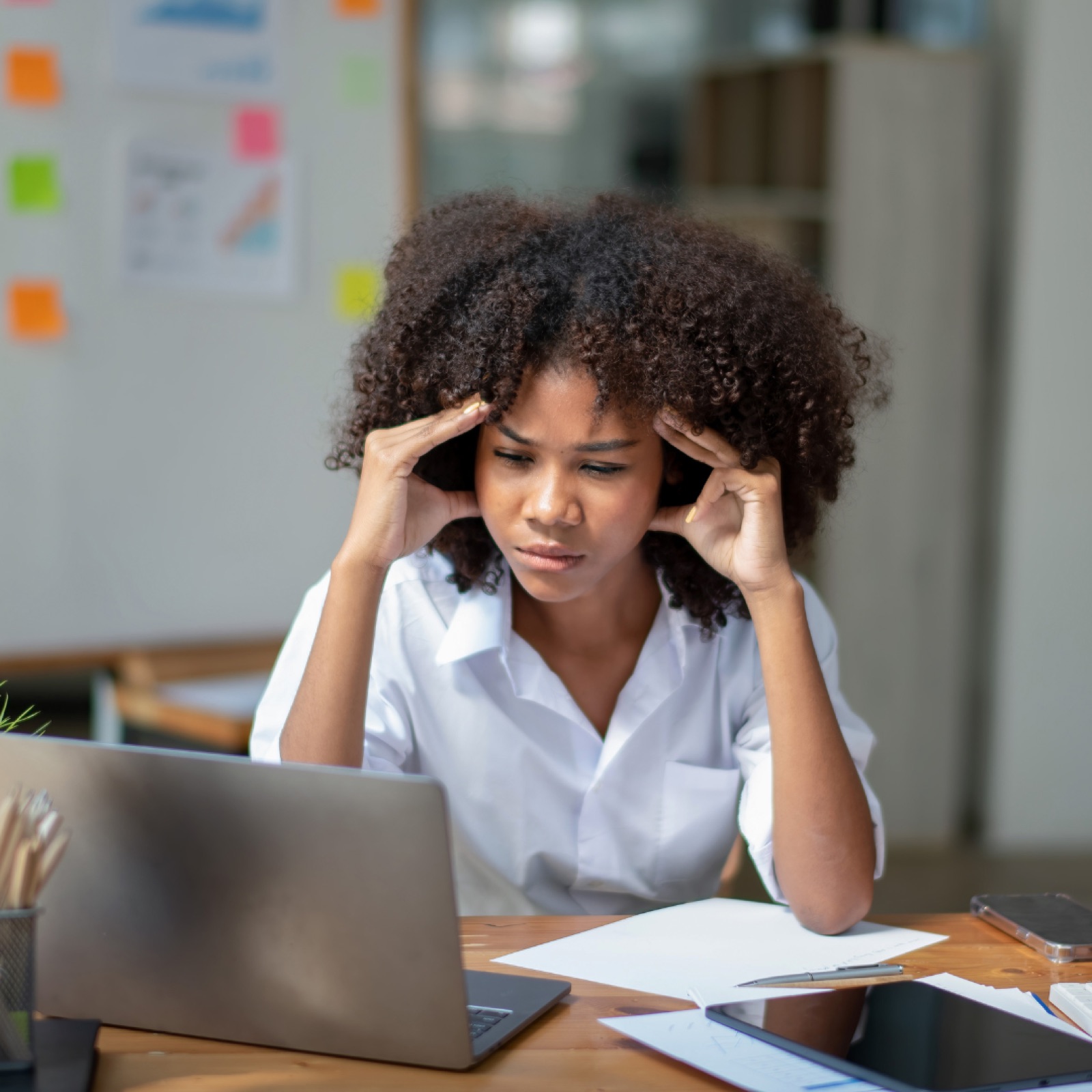 Woman in a white shirt, stressed, at a desk with a laptop and papers. Bright office with sticky notes on the wall. Focused mood.
