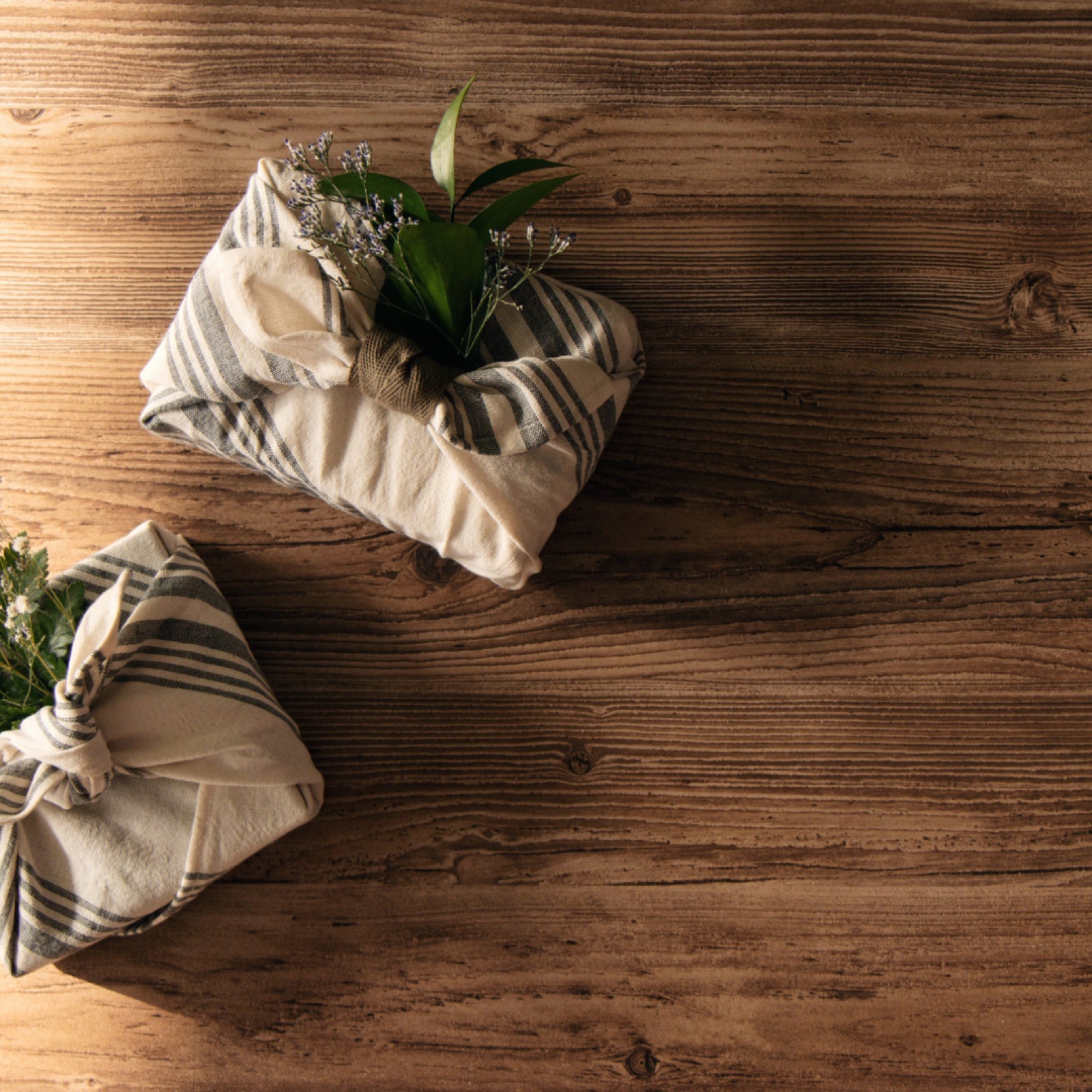 Fabric-wrapped gifts with green leaves and delicate flowers on a rustic wooden surface under warm lighting, creating a cozy, natural feel.