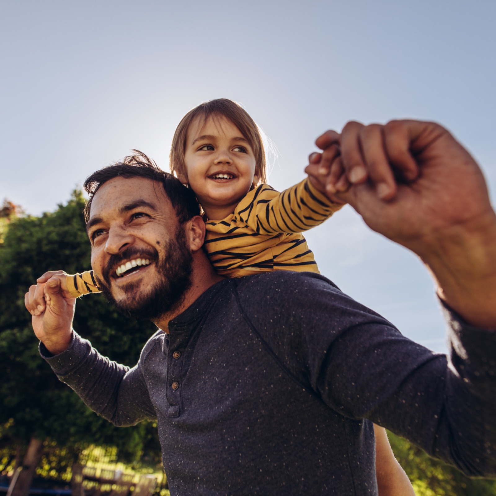 Man carries smiling child on shoulders outdoors. Child wears yellow striped shirt. Clear sky and green trees in background, cheerful mood.