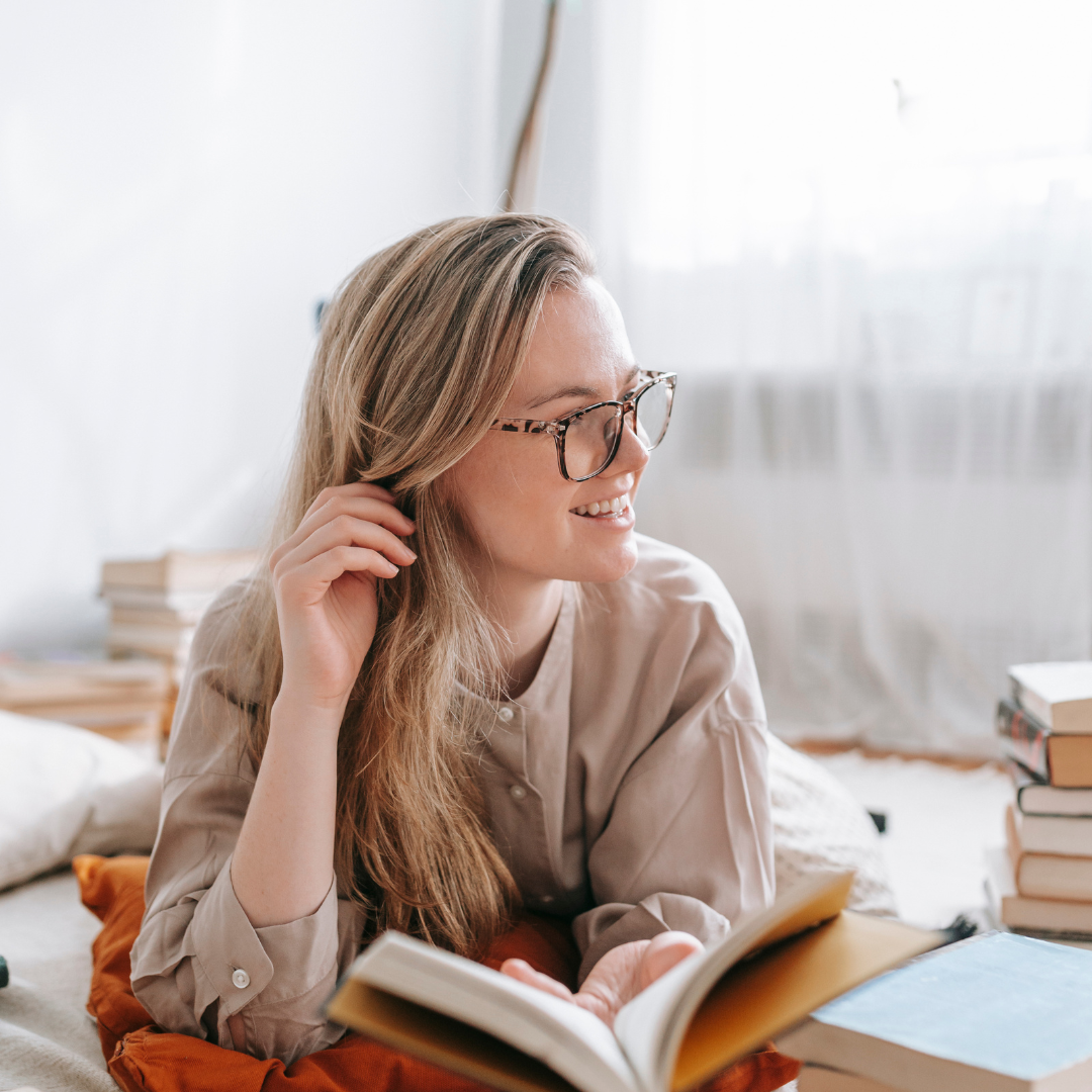 A person with glasses smiles while reading a book, lying on a bed. Stacks of books are around in a softly lit room with sheer curtains.