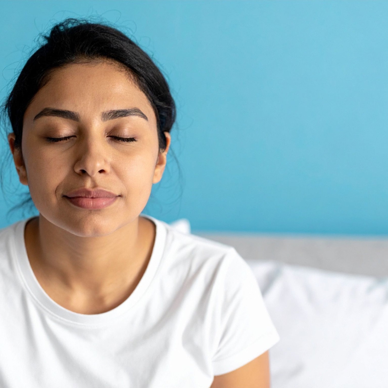 Woman in a white shirt meditates with closed eyes against a bright blue wall, conveying calmness and peace.