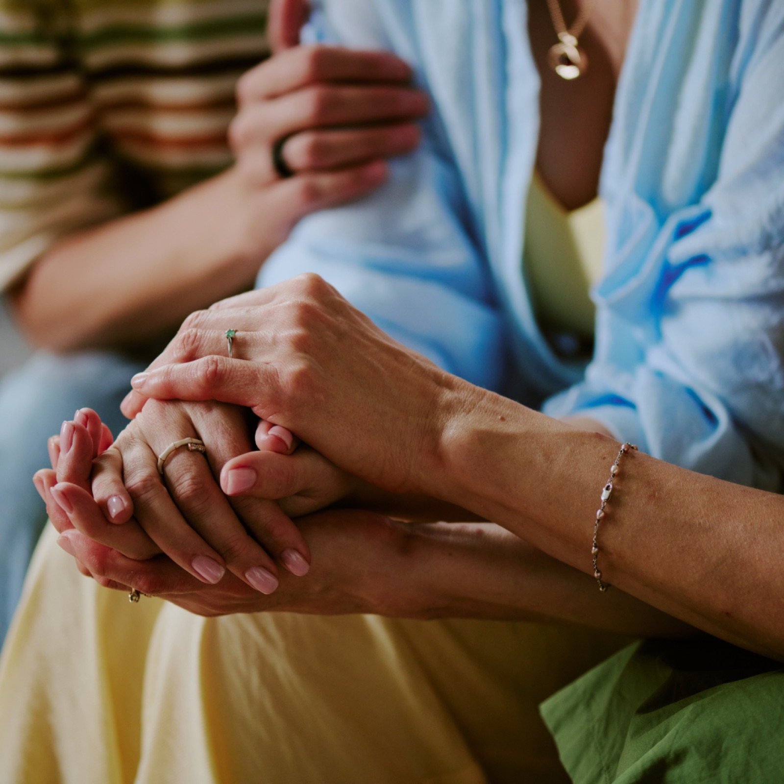 Two people are seated, holding hands in a comforting gesture. One wears a blue garment, and the mood is calm and supportive.
