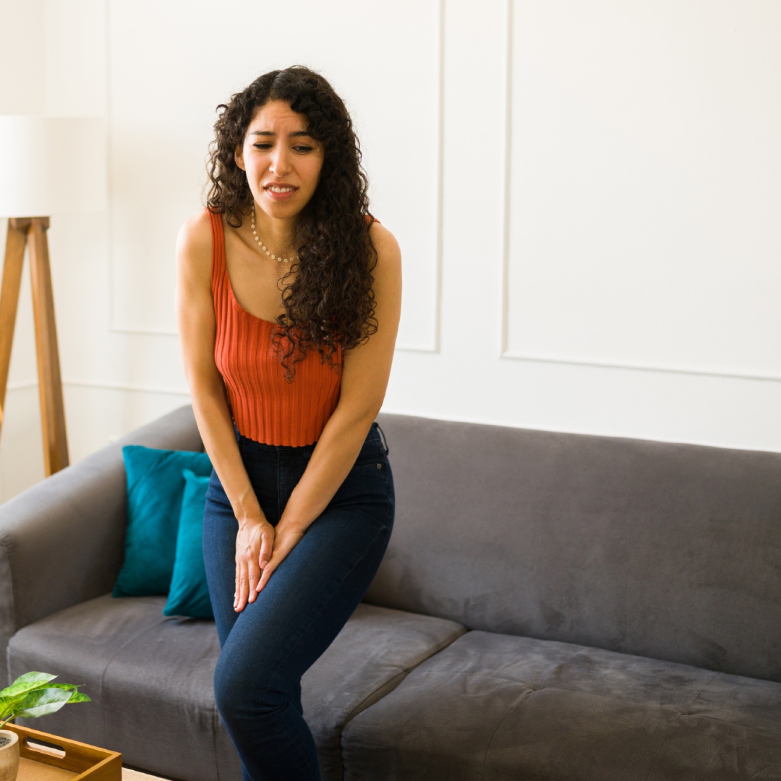 A woman in an orange top stands, looking uncomfortable, in a living room. She's in front of a gray sofa with teal pillows.