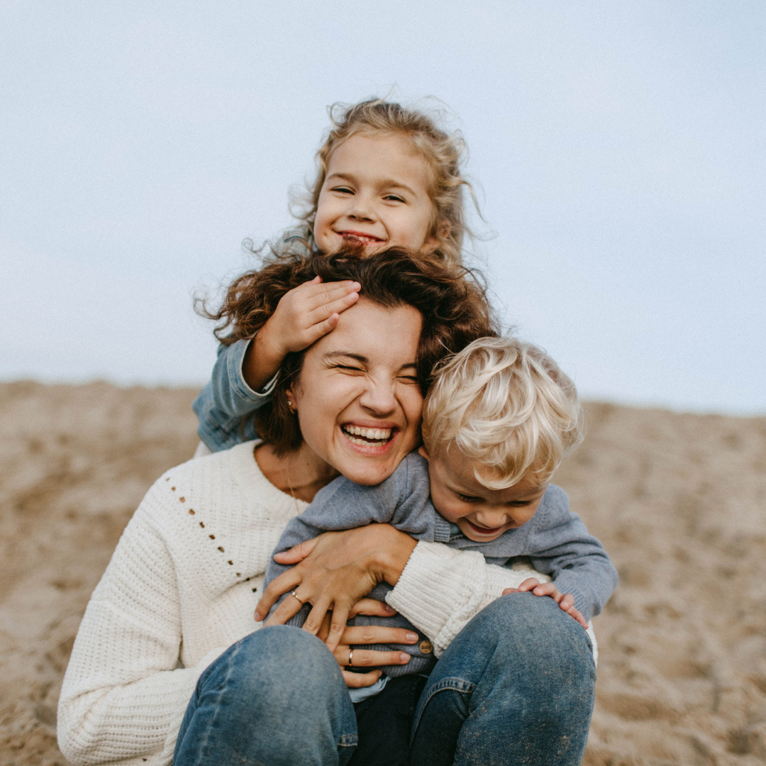 Woman joyfully hugging two children at a sandy beach. Bright sky, casual clothing, and playful mood. Kids smiling and laughing.