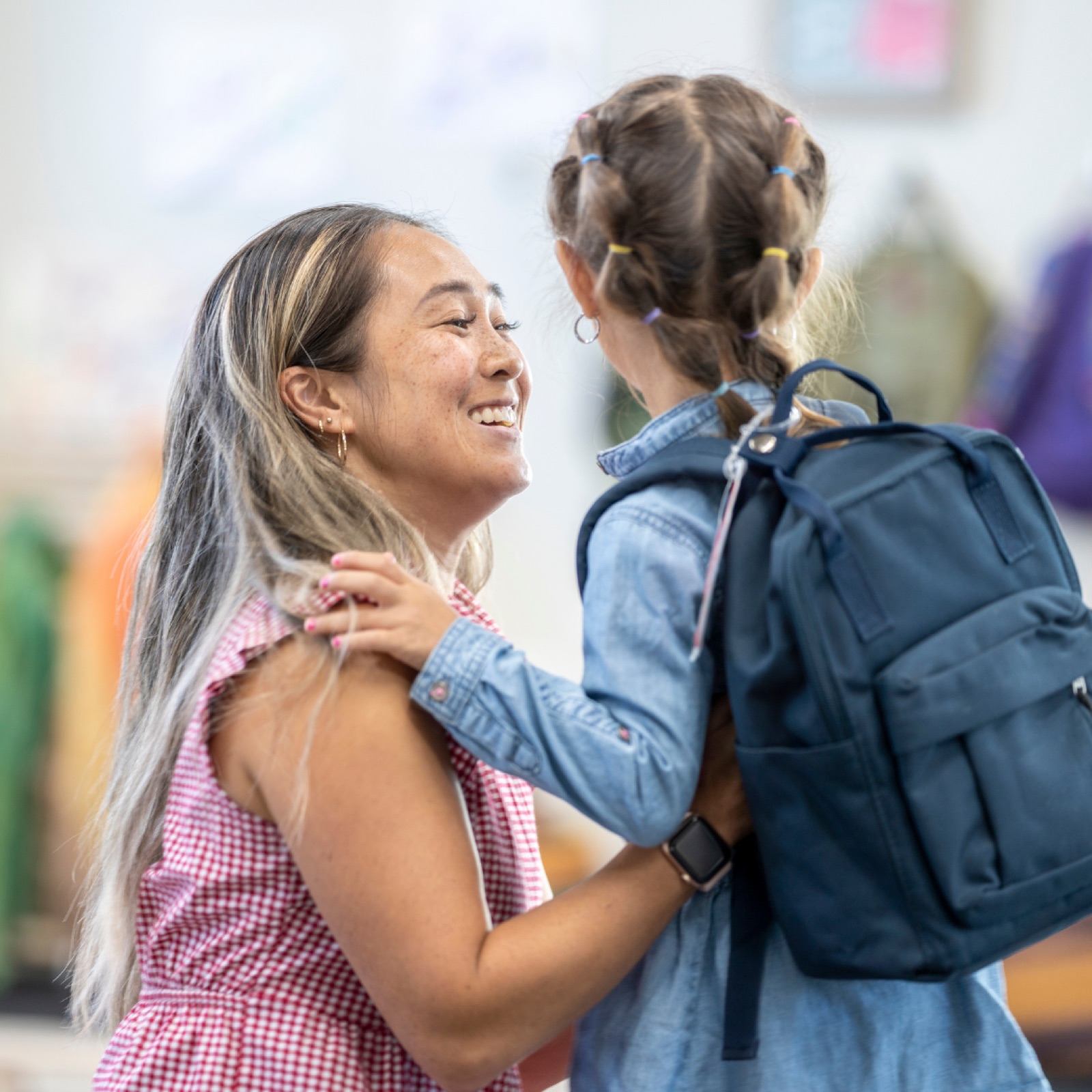 Woman smiles warmly at a child wearing a blue backpack. They're in a classroom setting with colorful decorations in the background.